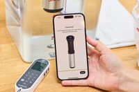 A hand holds a smartphone with the Anova Culinary Precision Cooker 3.0 setup confirmation displayed next to a sous vide container and a timer device