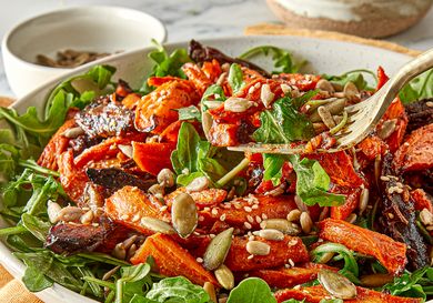 A roasted carrot and date salad with seeds on a bed of greens served in a wide bowl with a fork