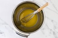 An All-Clad mixing bowl containing a whisk and a yellow liquid on a marble surface