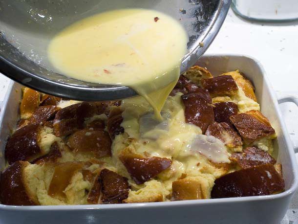 Pouring excess custard over soaked bread chunks in a baking dish.
