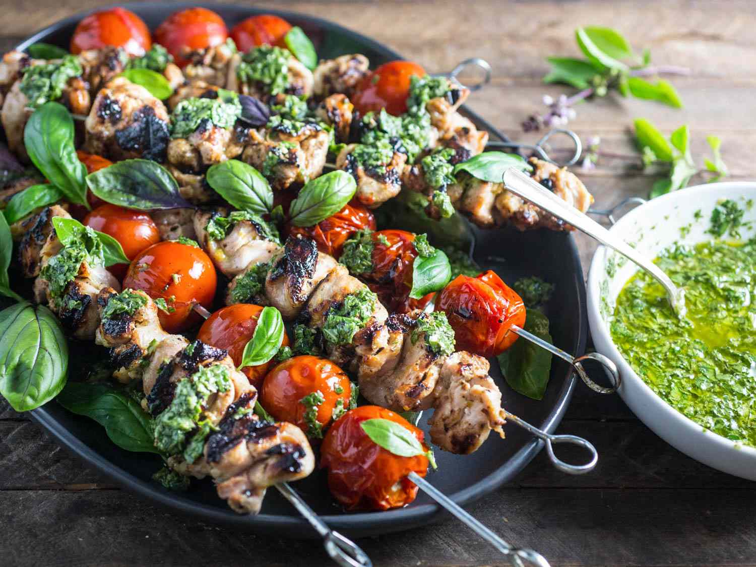 A black platter filled with chicken and cherry tomato skewers drizzled with chimichurri and garnished with fresh basil leaves. A bowl of chimichurri is next to the platter.