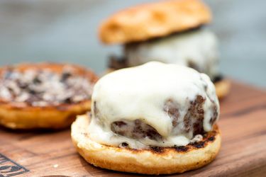 Profile view of a grilled black-olive burger, enrobed with melted Swiss, ready to be assembled with the top bun.