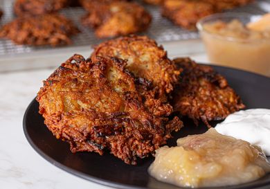 A plate of fried latkes served with applesauce and sour cream additional latkes cooling on a rack in the background