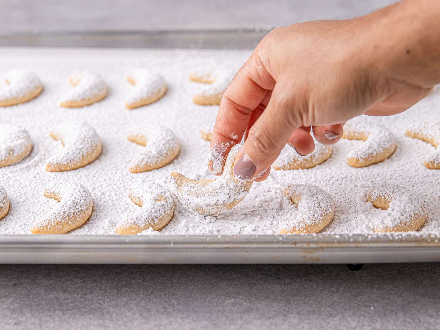 Hand sprinkling powdered sugar over crescent shaped cookies on a tray