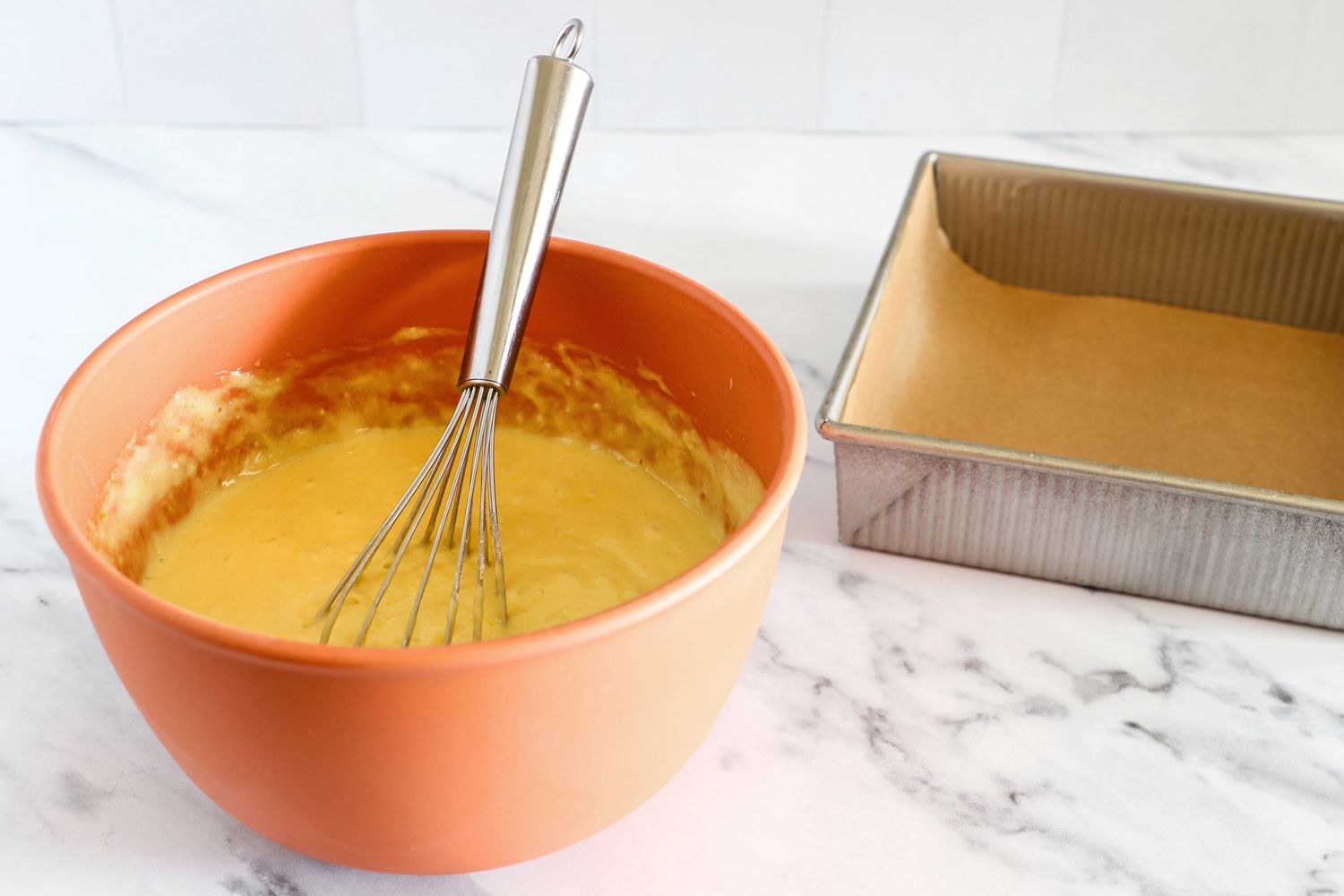 A Bamboozle mixing bowl with batter and a whisk next to a loaf pan lined with parchment paper on a counter