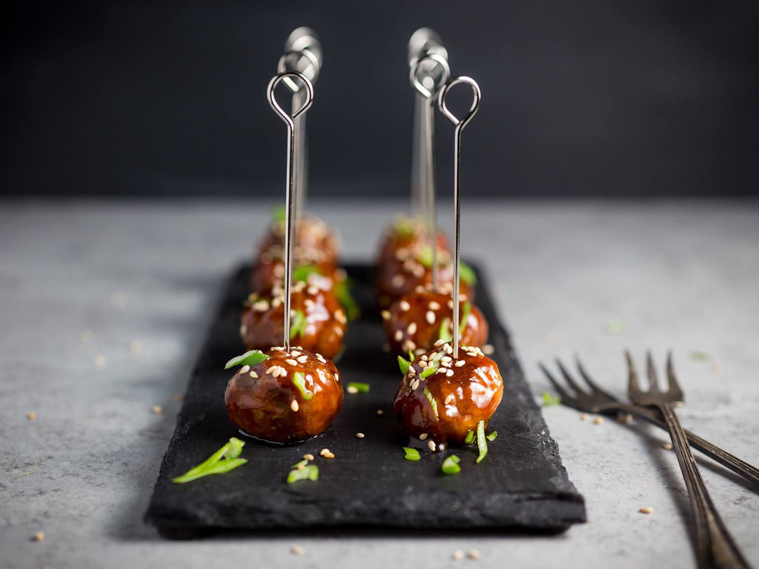 A dark slate tray with neatly lined up hoisin meatballs with fancy silver skewers, topped with sesame seeds and sliced scallions.