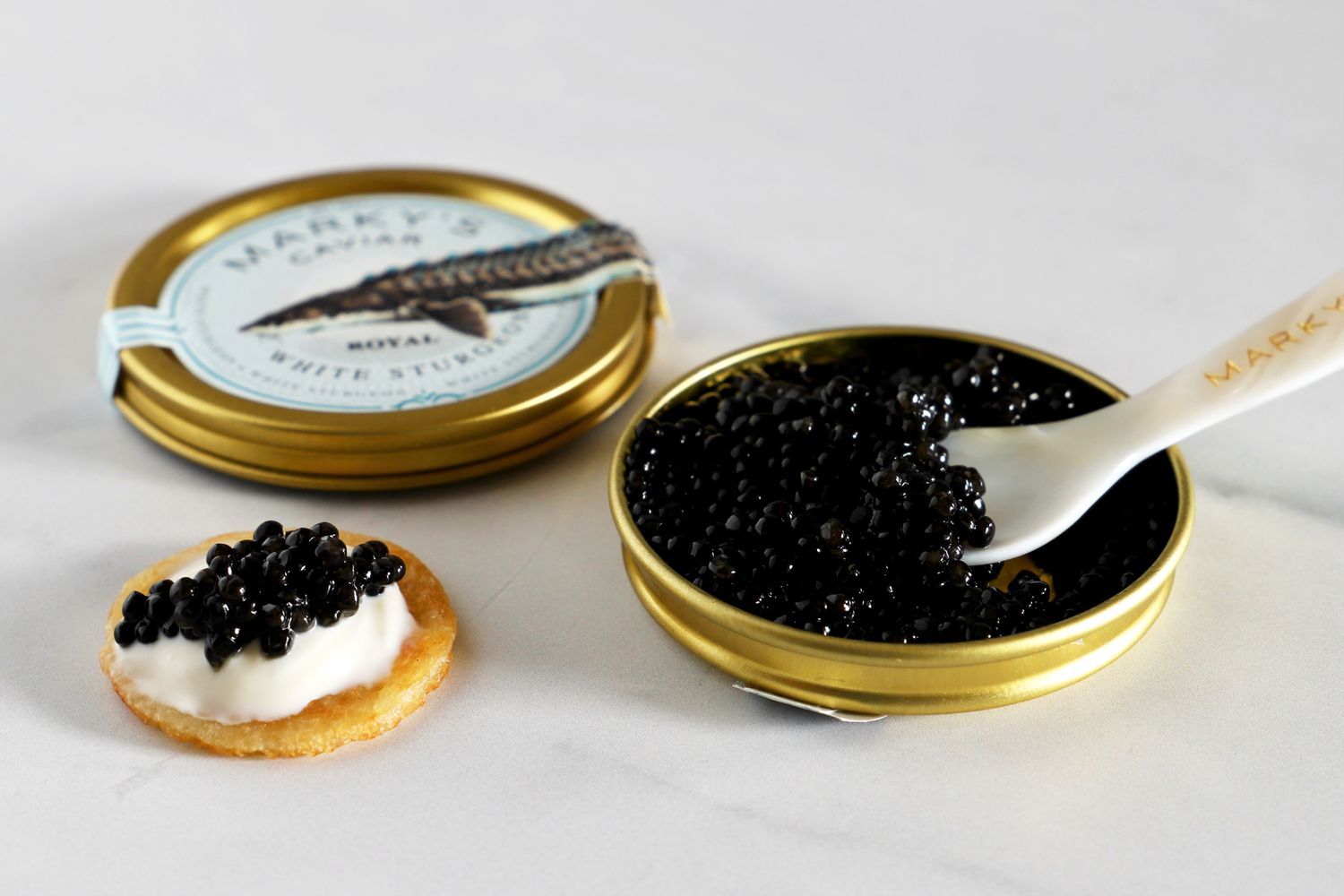 a tin of Marky's royal white sturgeon caviar next to a blini topped with crème fraîche and a scoop of caviar