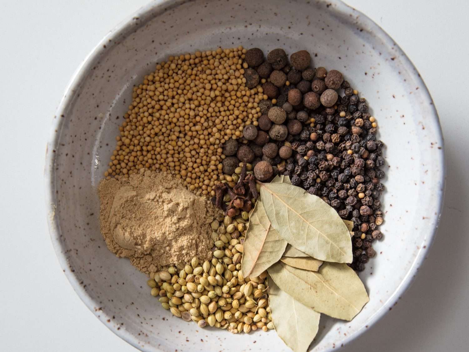 A bowl with ingredients for corned beef rub: mustard seed, allspice, black peppercorns, bay leaves, coriander, and ground ginger.