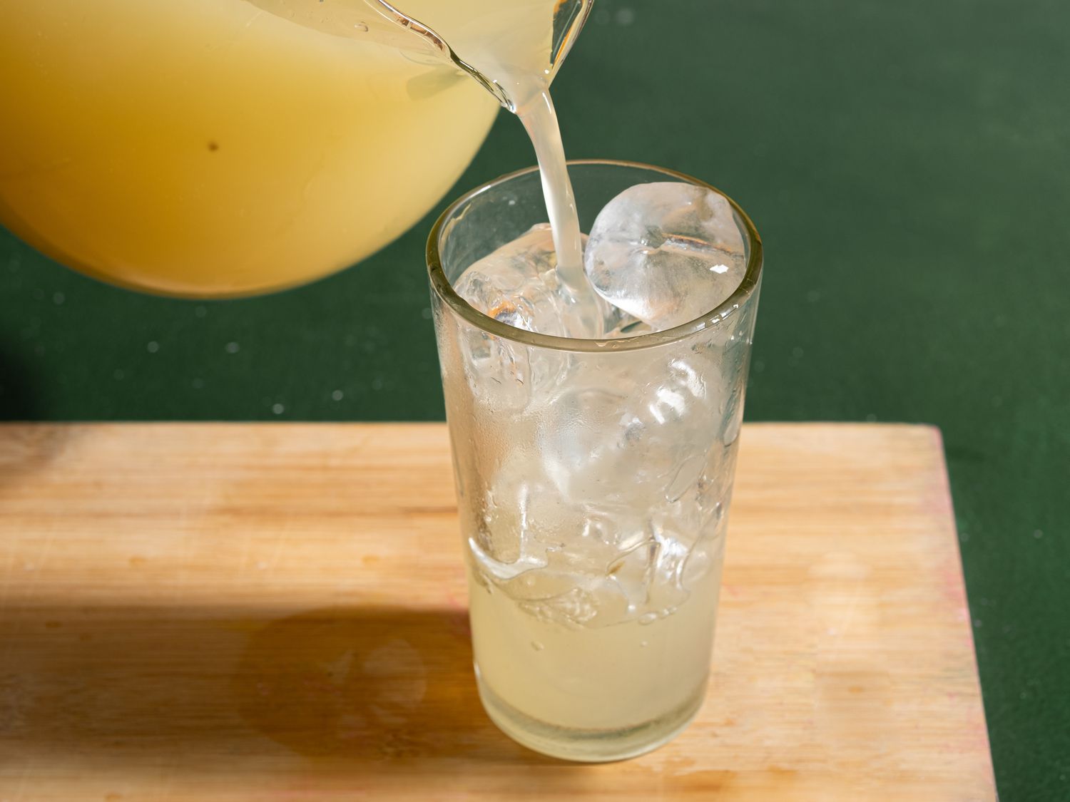 A jug pouring lemonade into a glass filled with ice on a wooden surface