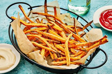 Basket of sweet potato fries on a blue surface, with small dishes of ketchup and aoli sause, and a bottle of water