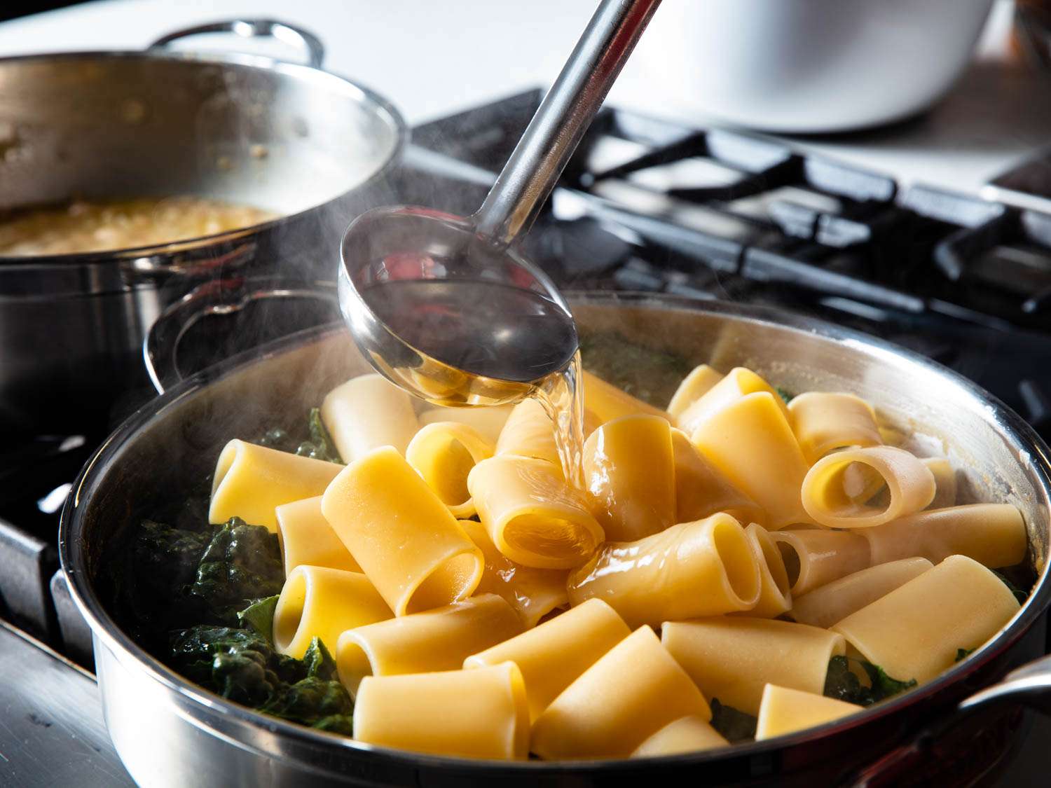 A ladle pouring water onto pasta and greens in a pan. 