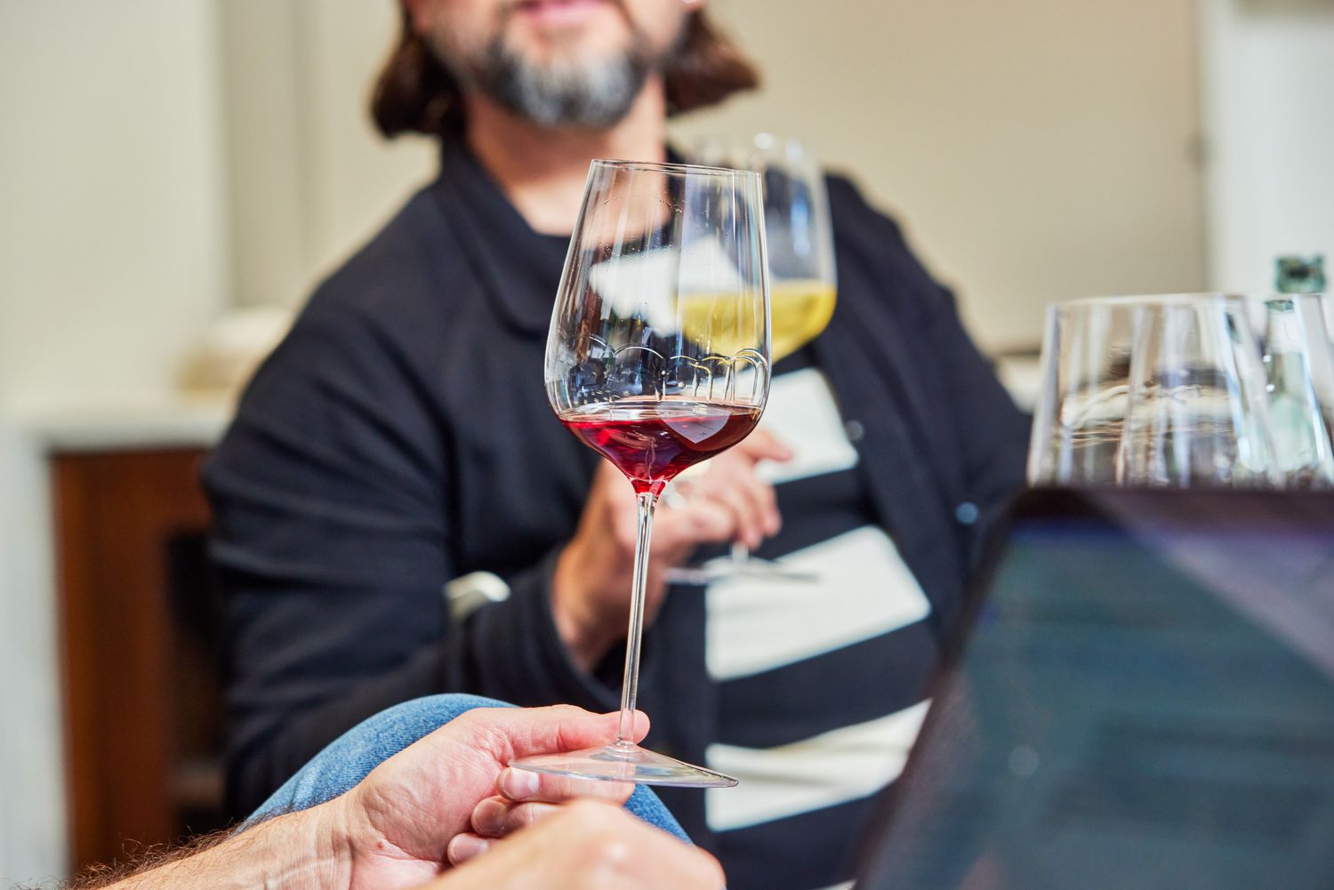 A hand holding a glass from Glasvin Universal Wine Glasses, Set of 2, with red wine in it and a person in background