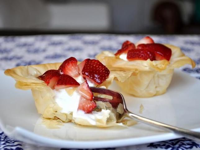 Two Greek yogurt and strawberry phyllo cups, served on a small plate. One has been partially demolished with a fork to reveal the yogurt filling.