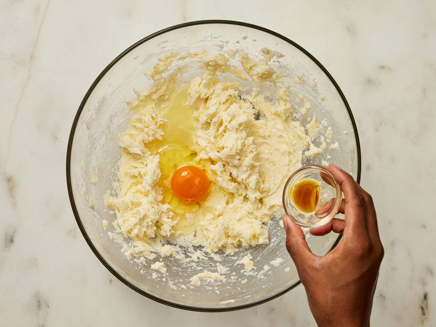 Hand pouring small dish of vanilla and an egg to cookie mixture in bowl 