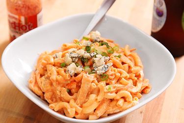 Closeup of buffalo chicken mac and cheese, served in a bowl with a sprinkle of crumbled blue cheese. A bottle of Frank's Red Hot sauce lurks in the background.