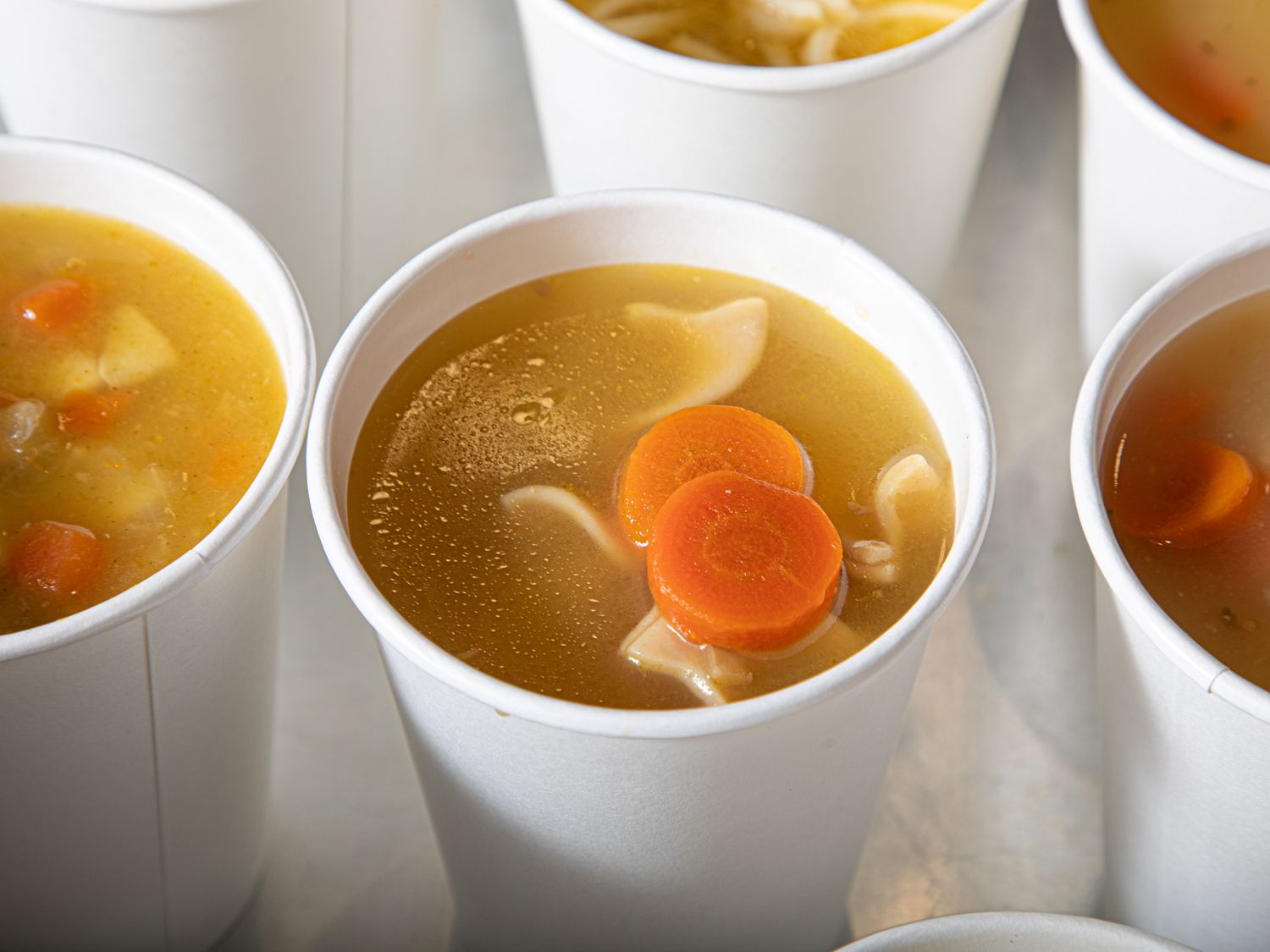Array of cups containing chicken noodle soup with carrots and noodles visible