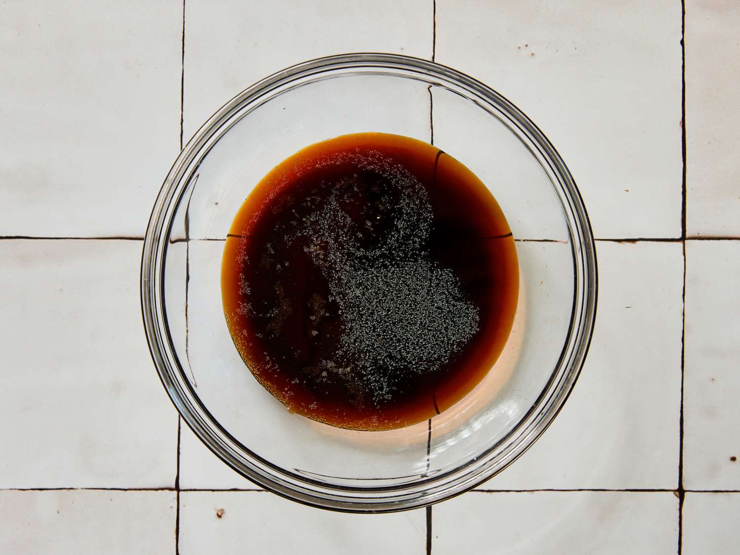 A glass bowl containing a dark liquid mixture on a white tiled surface