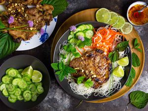 Overhead shot of a bowl of vermicelli noodles topped with grilled pork chops, carrot-daikon pickles, cucumbers, perilla/shiso, mint, cilantro, crushed peanuts, and a few stray flowering chives. A small bowl of nuoc cham is close at hand, as well as a perilla-leaf-lined plate piled with more of the grilled pork chops and a bowl of sliced cucumbers and lime wedges.