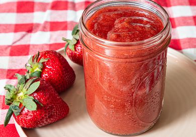 Glass jar of jam with whole strawberries on a plate and checkered tablecloth