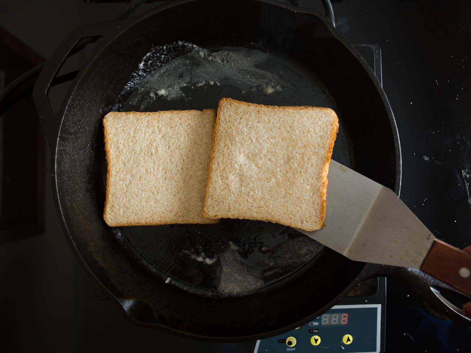Two slices of bread inside of a buttered cast iron skillet, with one piece being lifted by a broad, flat spatula.