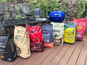Seven bags of lump charcoal lined up in front of a kettle grill on a deck.