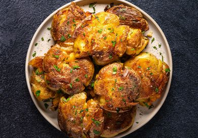 Golden brown crispy smashed potatoes on an oval white plate on a dark textured background.