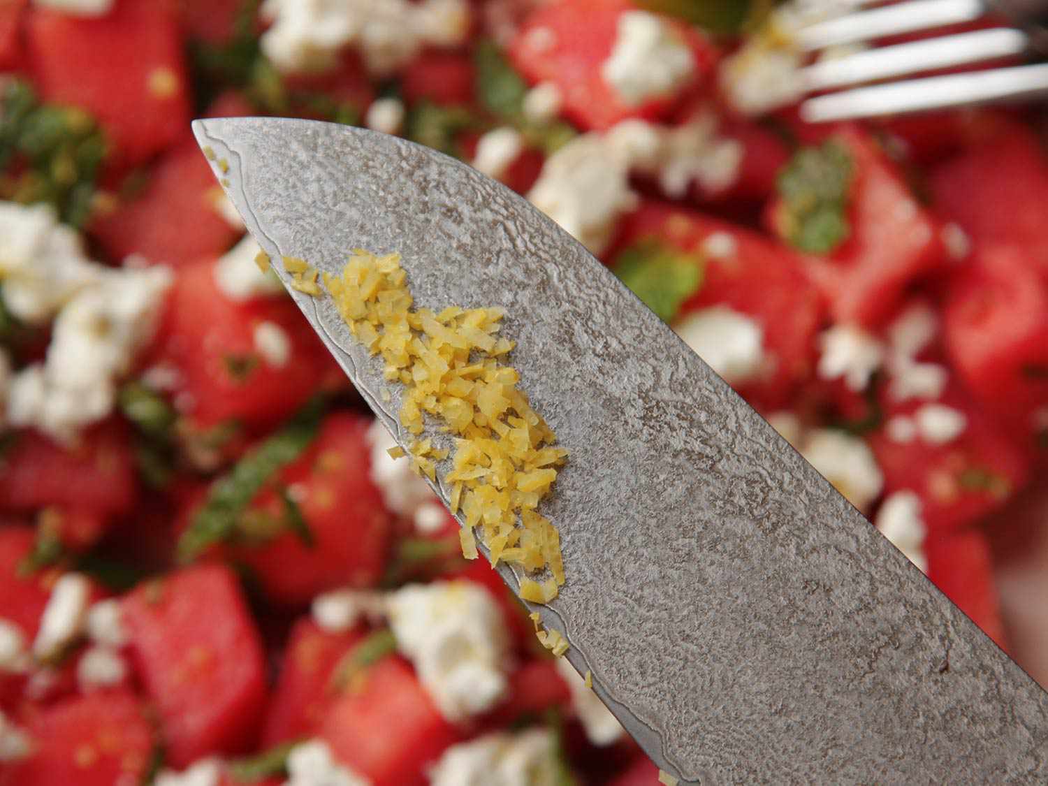 Chopped lemon zest on the blade of a chef's knife, with watermelon, feta, and mint salad in the background.
