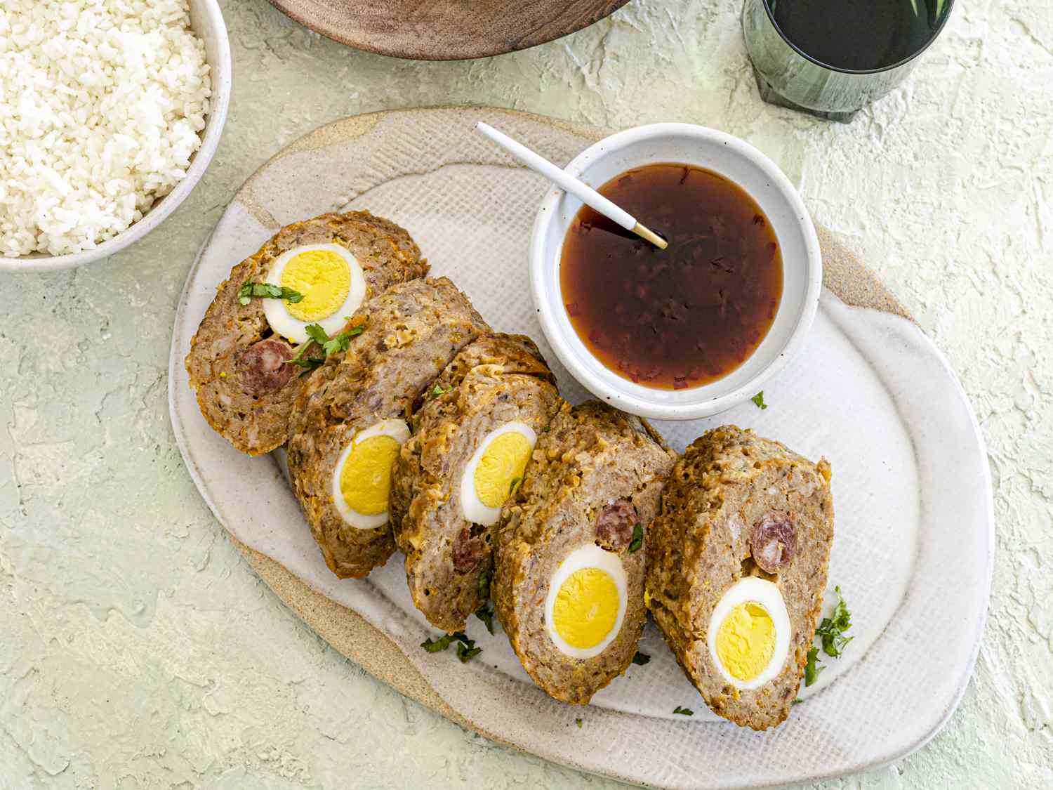Cut and plated embutido on a white and tan plate with sauce in a small bowl next to it and a serving of rice.