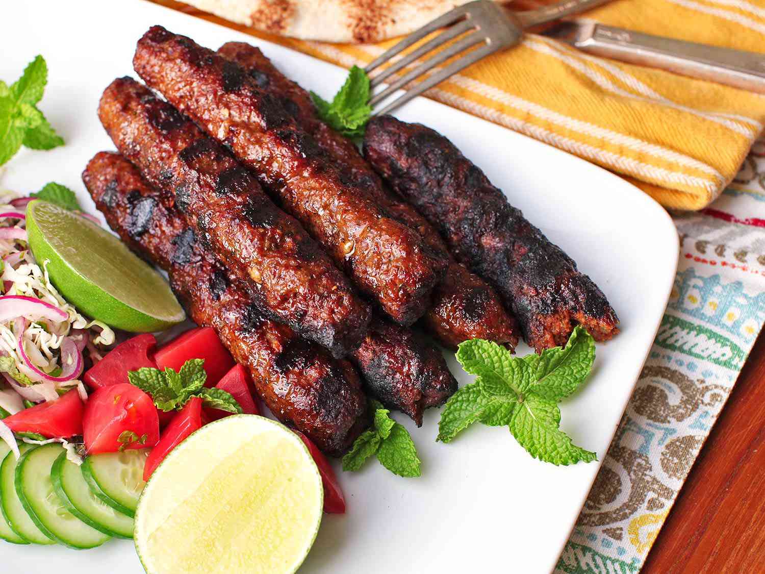 A platter of seekh kebabs with red onion and cabbage salad, lime wedges, and fresh herbs. 