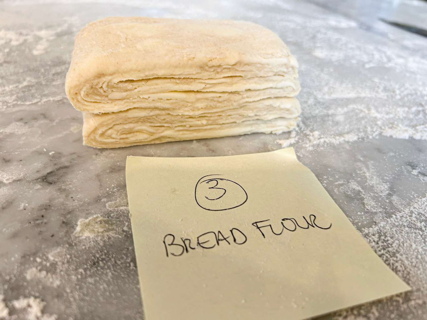 A stack of folded dough with a note labeled 3 Bread Flour on a floured surface