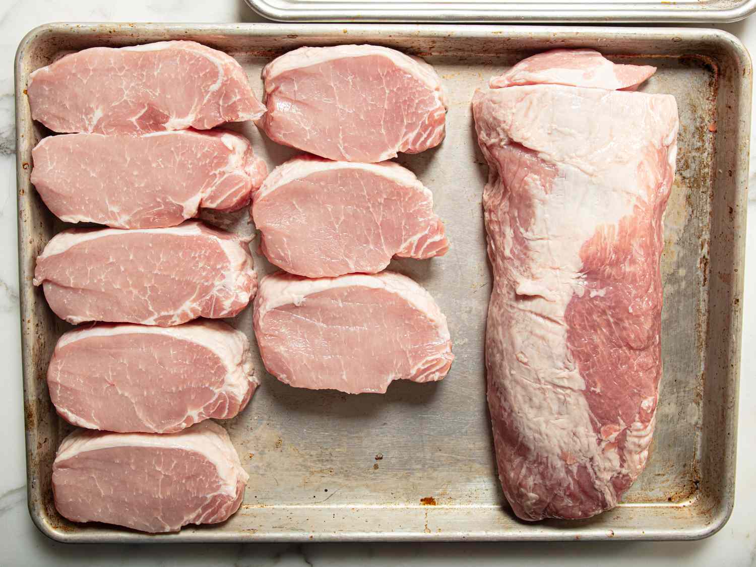 Cuts of raw pork loin next to a whole pork tenderloin on a baking sheet