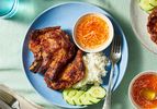 Pork chops over steamed rice white on a plate with sliced cucumbers and a ramekin of sauce