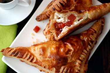 Overhead view of guava and cream cheese pastelitos, served on a square white plate with a cup of coffee nearby. One of the pies has been bitten, revealing the filling to the camera.