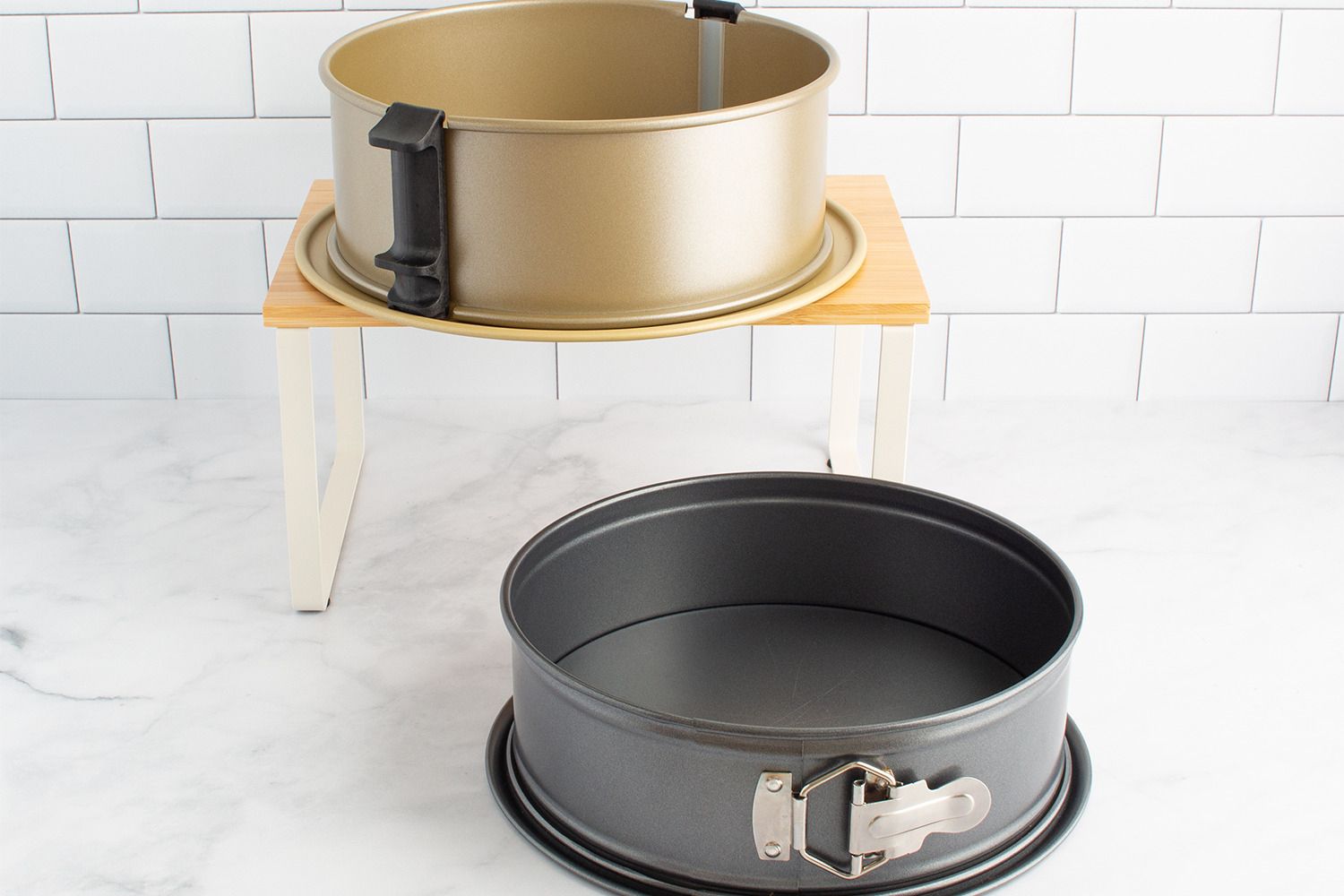 Two springform pans one black and one bronze displayed on a light surface with one elevated on a small wooden stand