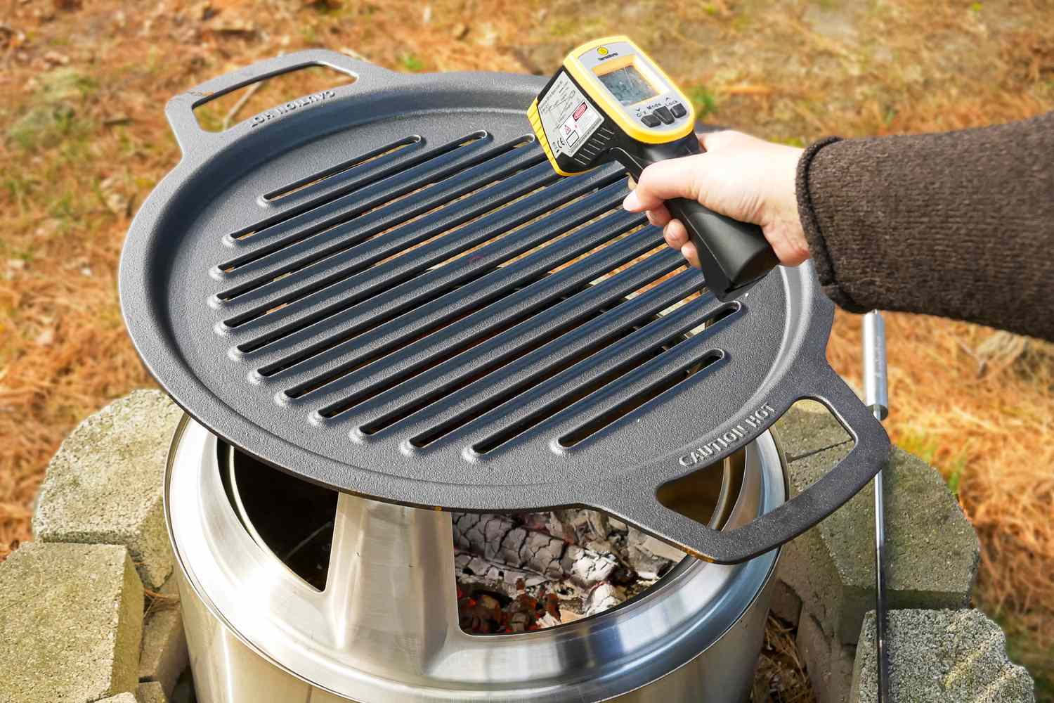 A hand holding a infrared thermometer measuring the temperature of a grill atop a metal outdoor stove with glowing embers beneath