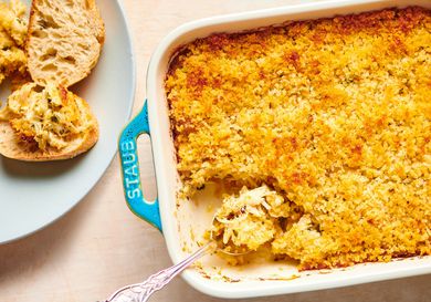 A blue baking dish holding golden brown Maryland Crab Imperial. The bottom left corner of the baking dish shows a metal spoon holding a spoonful of the crab, while on the lefthand side of the image is a light blue ceramic plate holding small pieces of bread, one of which has been topped with the crab.