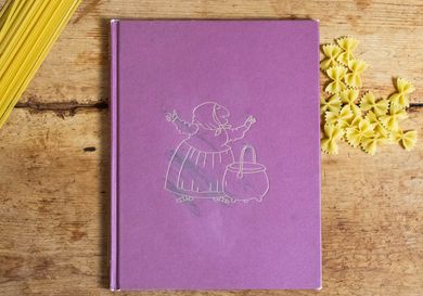 A copy of the book Strega Nona by Tomie dePaola, on a wood tabletop with dried pasta. .