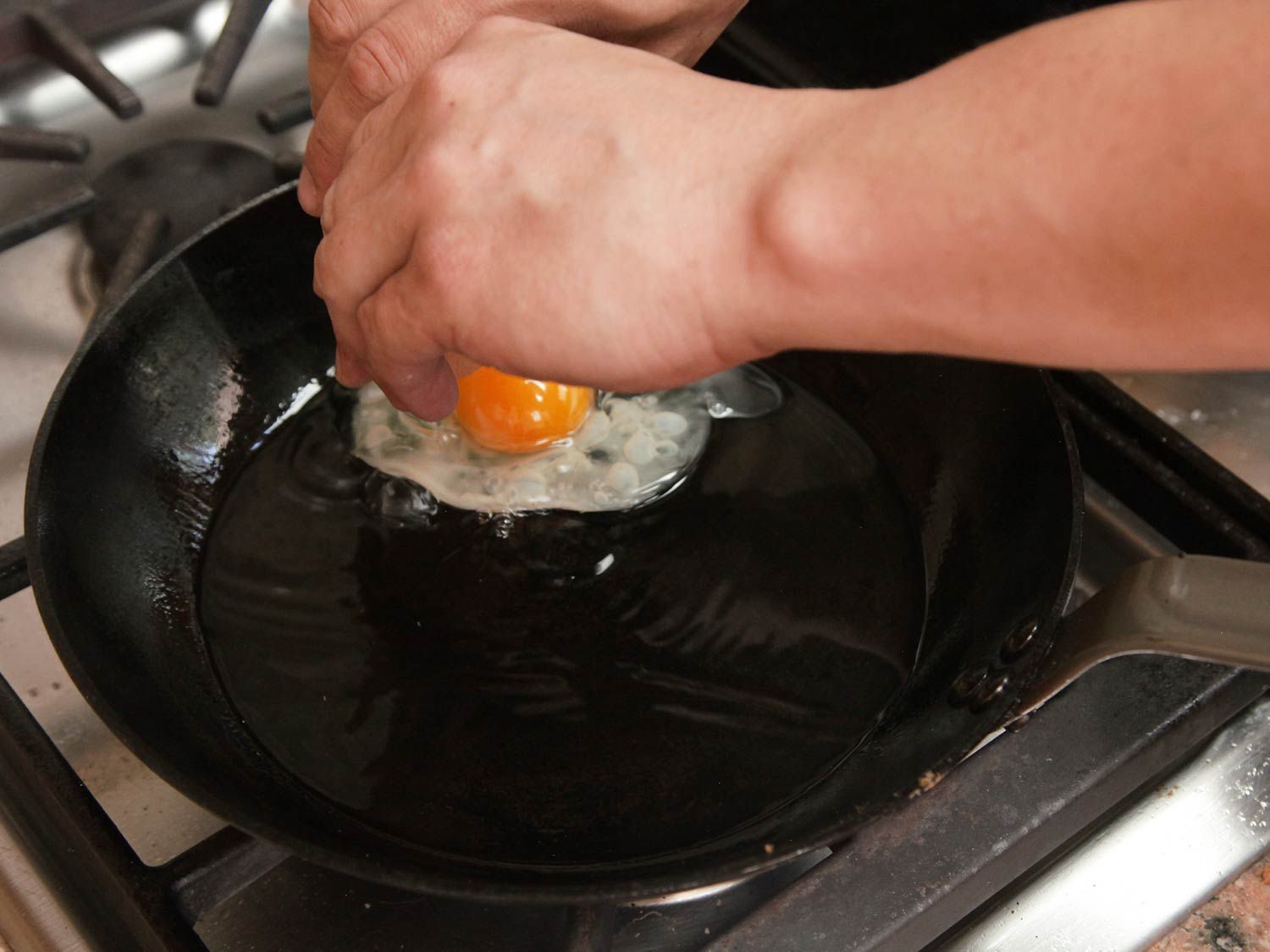 Two hands cracking an egg into hot oil in a skillet.