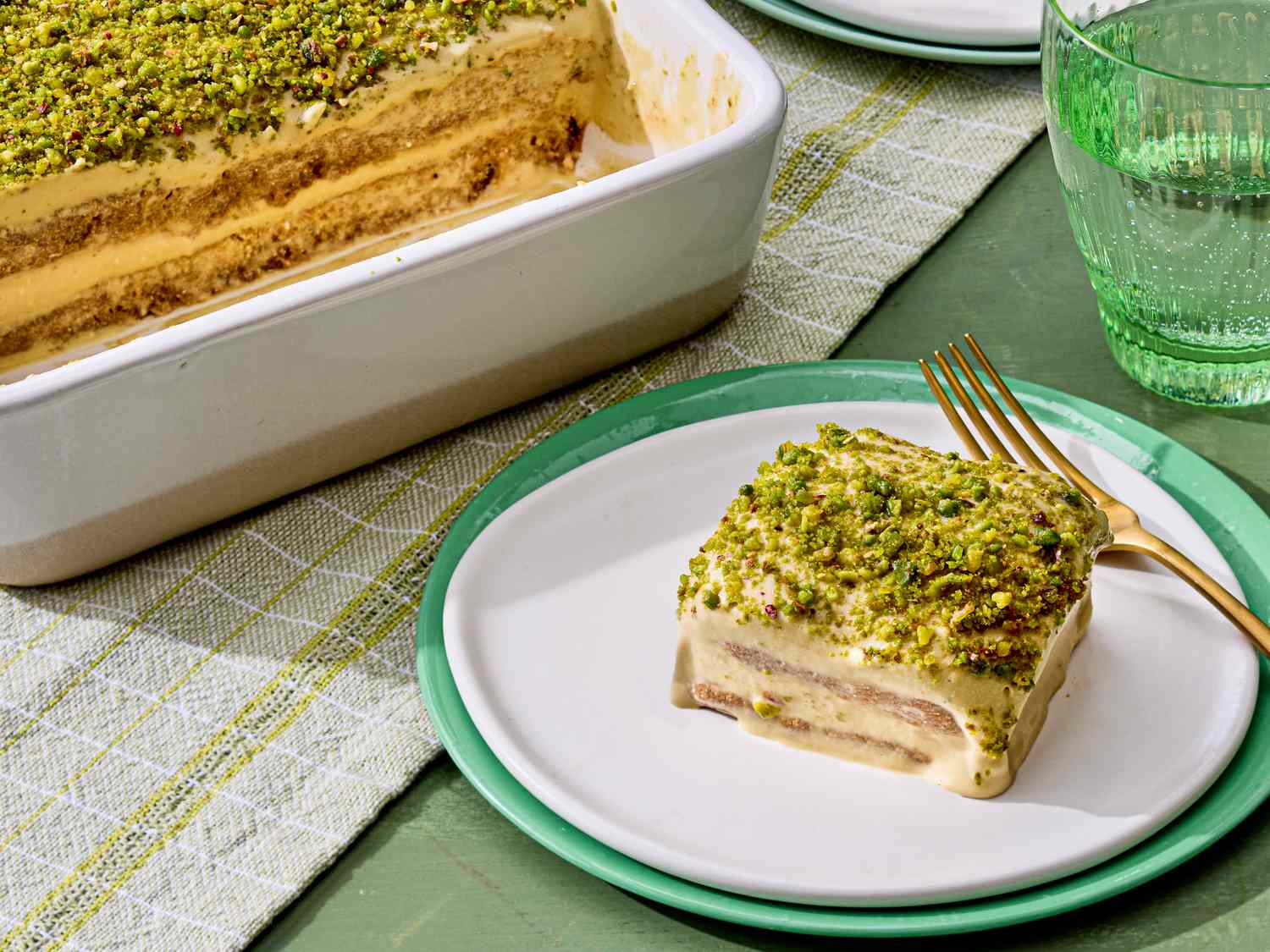 A piece of pistachio tiramisu served on a plate next to the main dish tray