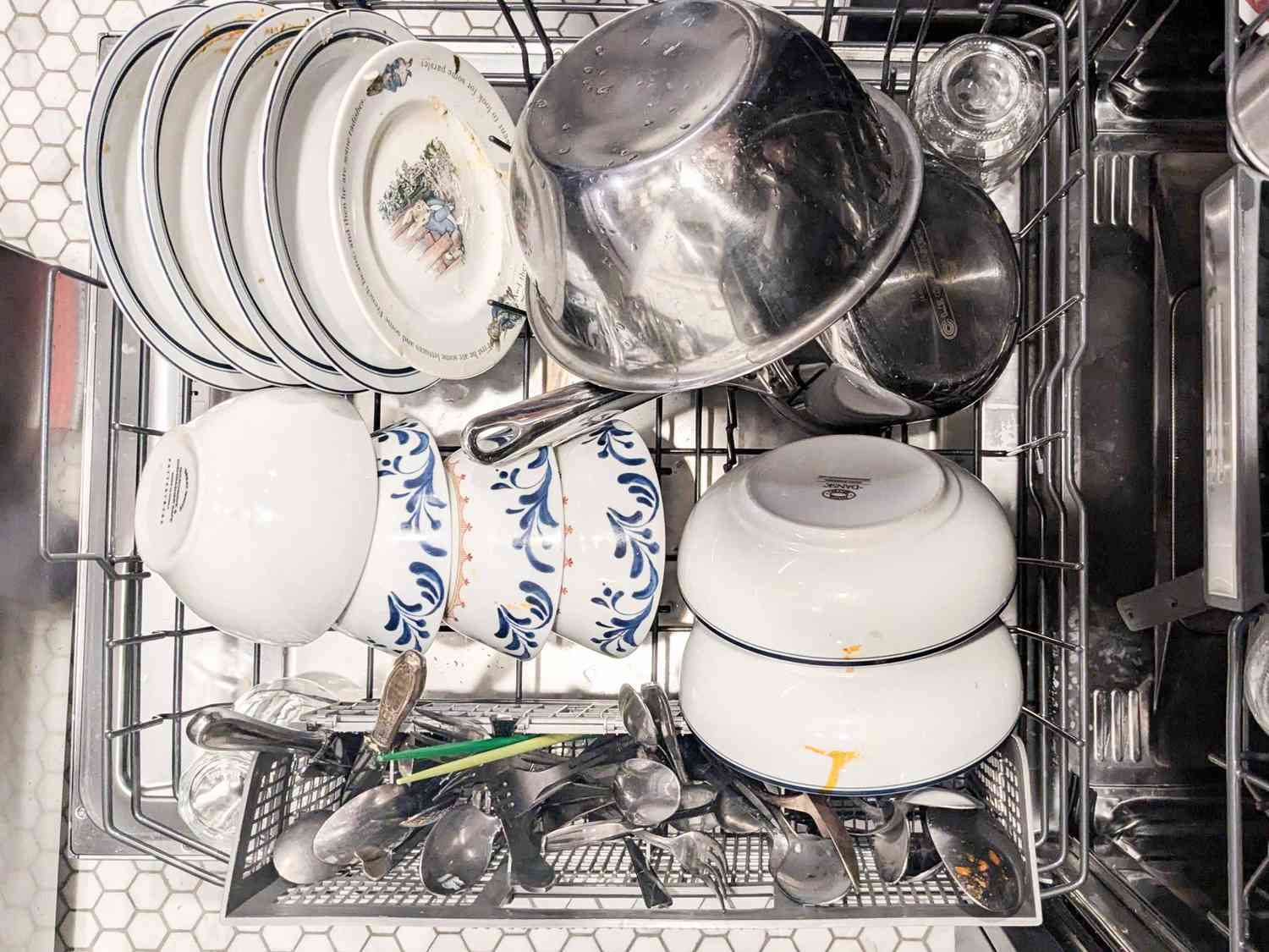 A bottom rack of a dishwasher neatly loaded with plates, bowls, mixing bowls, a pot, and silverware.
