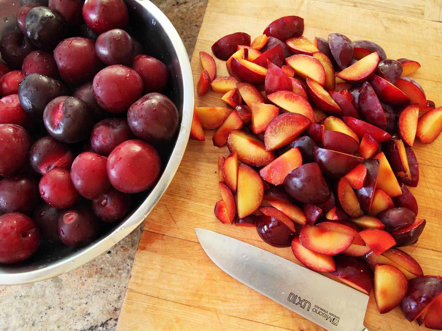 Sliced plum fruit on a wooden board next to a chef's knife, with a large metal bowl of whole plums nearby.