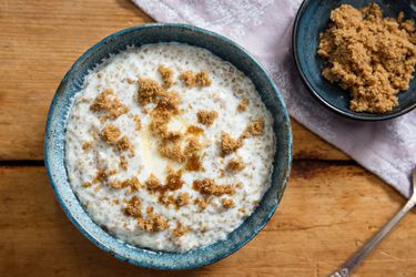 Overhead view of a blue ceramic bowl filled with creamy Irish-style oatmeal with brown sugar. A small bowl of brown sugar is nearby.