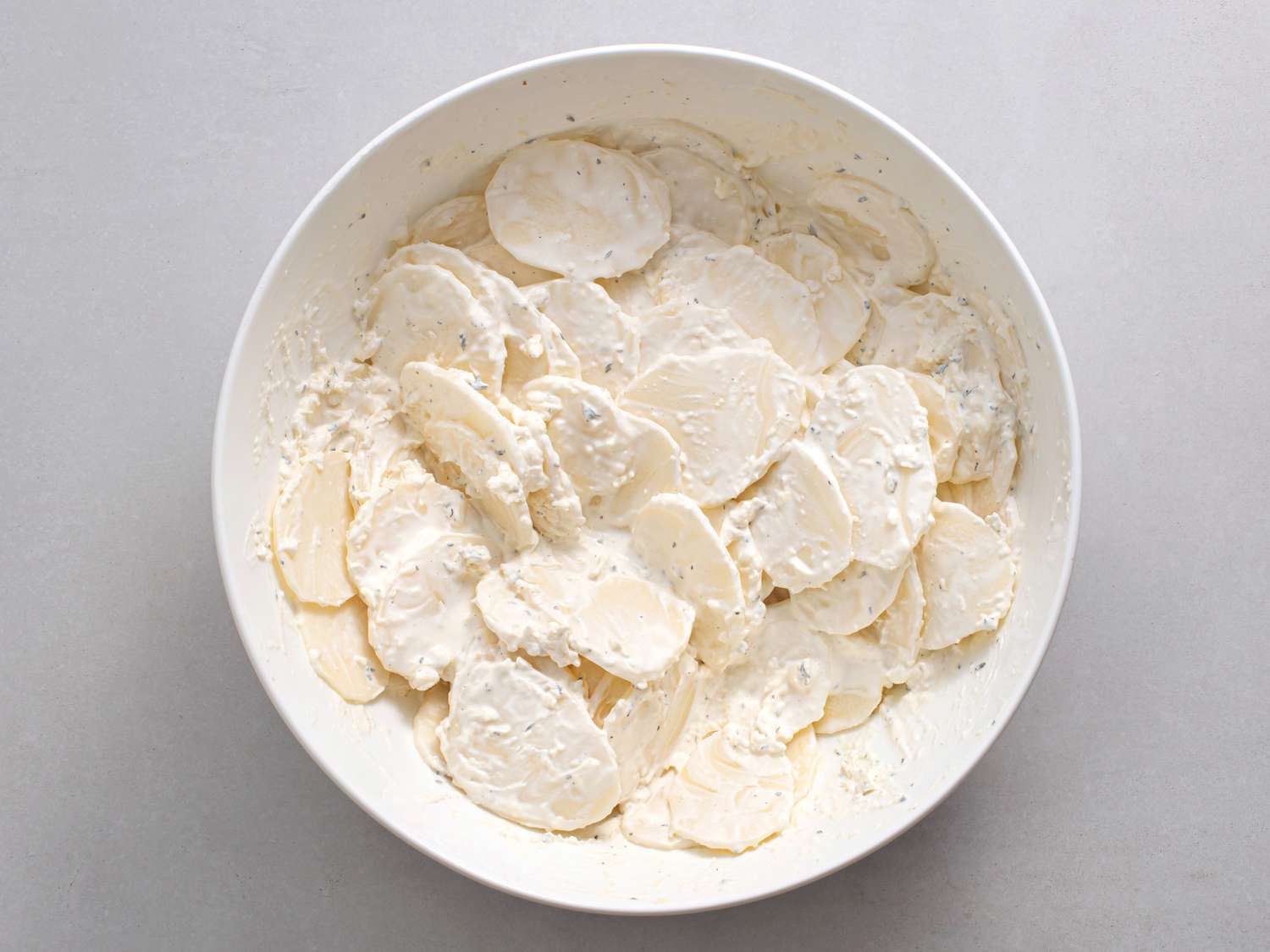 A white porcelain bowl filled with sliced potatoes mixed with cream, thyme, garlic, and cheese.