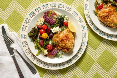 A plate of roasted chicken with vegetables and feta on a patterned plate on a green background additional plate and utensils visible