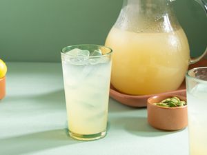 A glass of lemonade with ice cubes beside a pitcher containing more lemonade, next to small bowls of cardamom and lemons