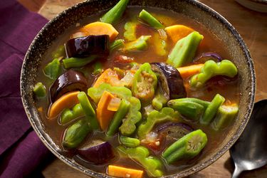 Bowl of pinakbet stew on a wooden surface, with a purple napkin to the left and a spoon to the right 