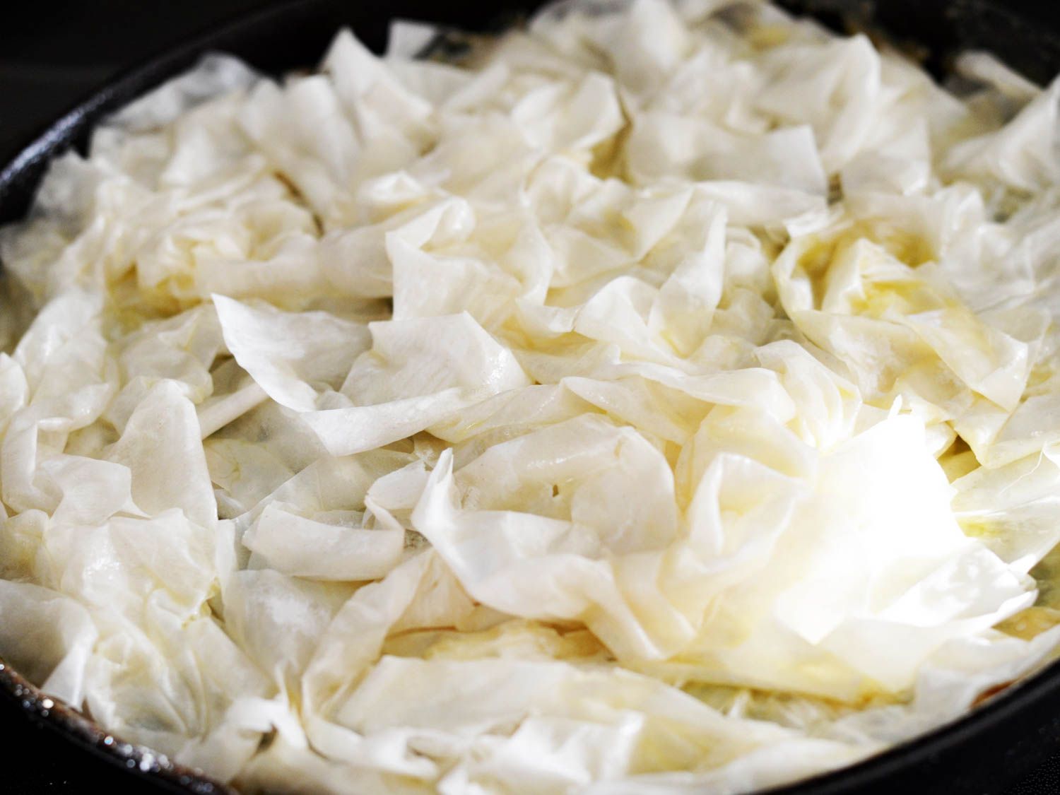 Crinkled-up, butter-brushed phyllo dough, uncooked, on top of chicken spinach filling in a skillet.