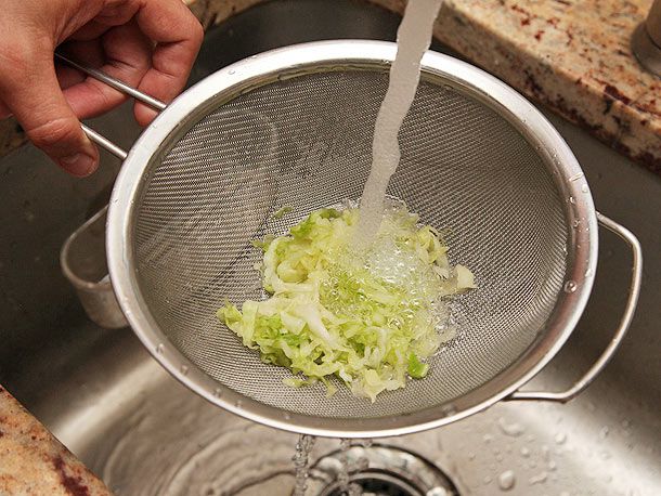 Hand holding fine mesh strainer of salted cabbage shreds in sink under running water