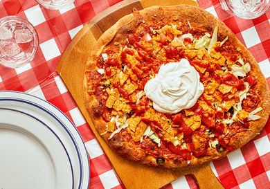 Taco pizza with sour cream on a wooden board, set on a red-checkered tablecloth, plates, and glasses nearby.