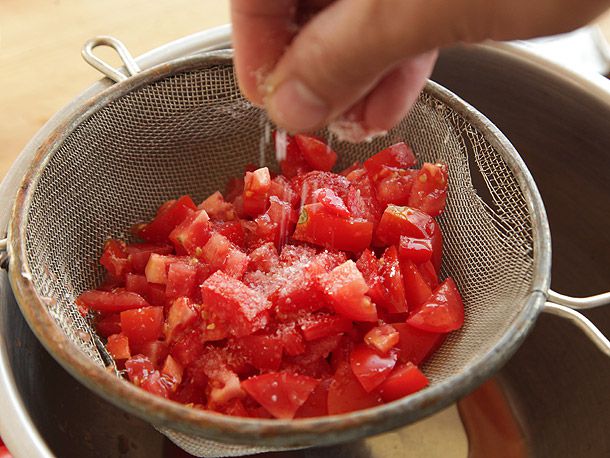 Hand sprinkling salt over pile of chopped tomatoes in fine-mesh strainer